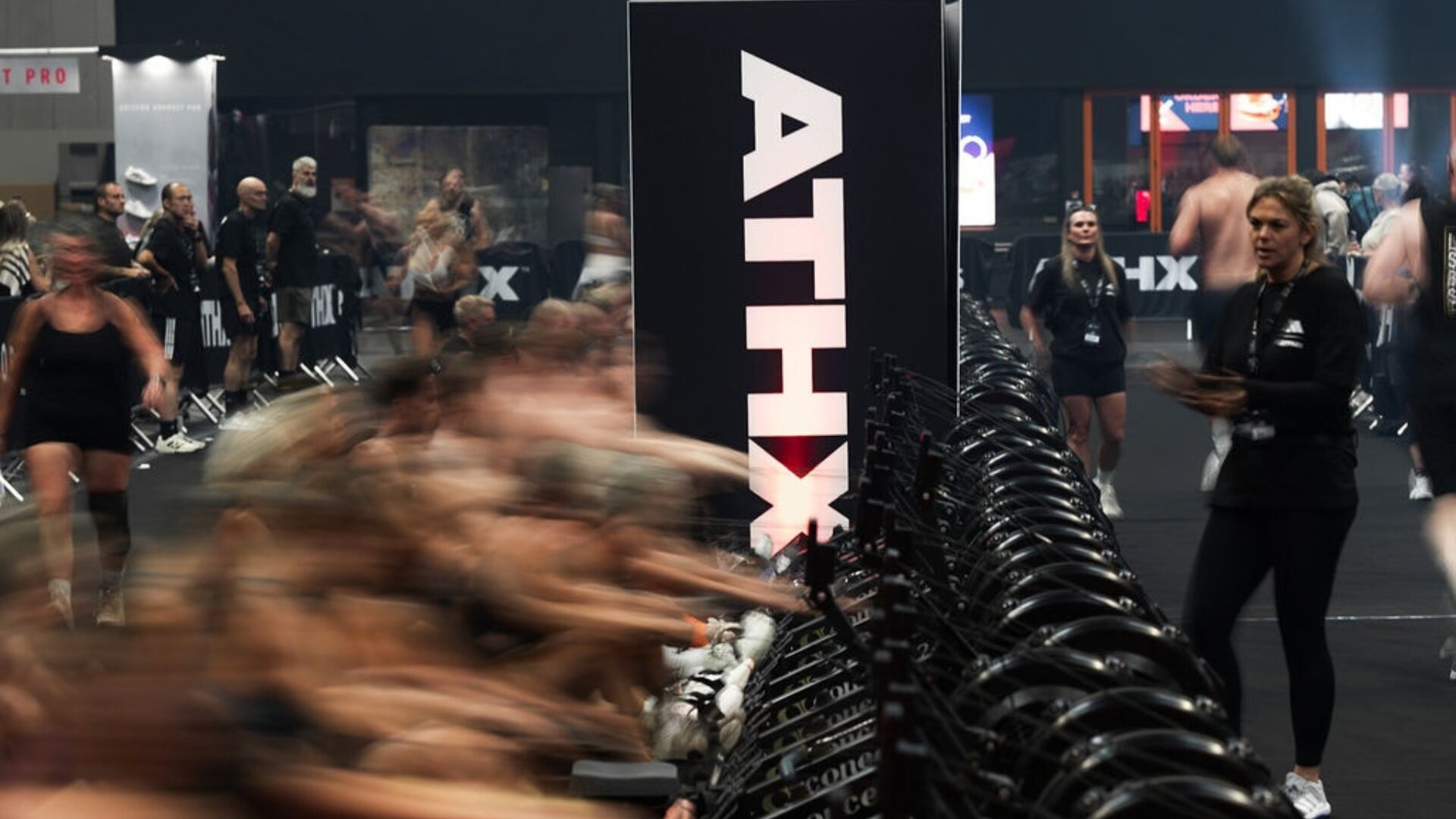 Athletes race on a long line of rowing machines beside a giant ATHX sign as staff look on