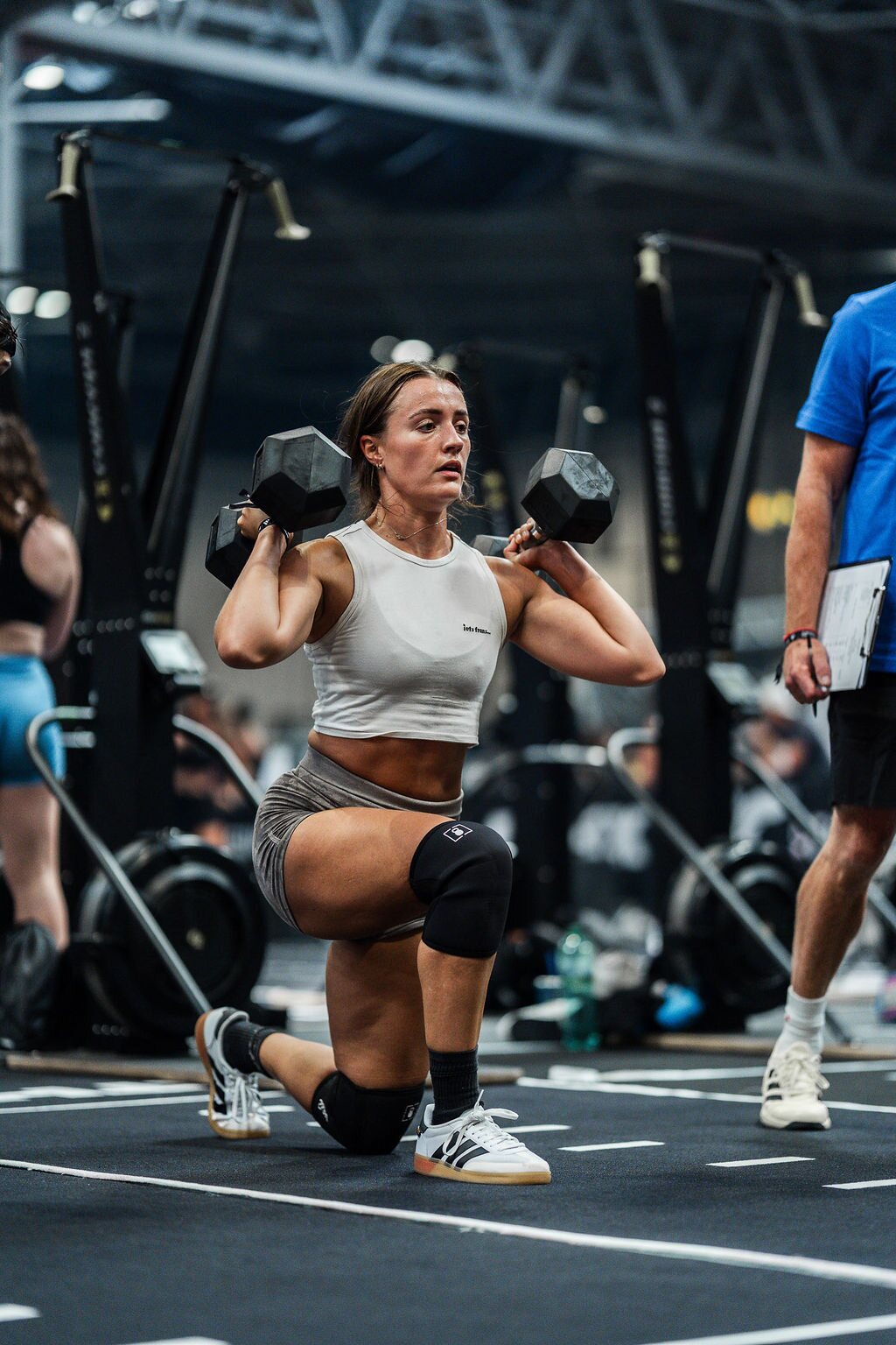 An athlete lunges with two dumbbells held at her shoulders as a judge watches